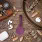 A purple leather luggage tag or phone strap accessory is displayed on a rustic wooden surface surrounded by styled props including a brown leather case, white wireless earbuds, a white charging case, dried flowers, eucalyptus leaves, and a large brass tray.