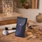 A navy blue leatherette pouch with a silver snap button, standing on a rustic wooden table surrounded by makeup brushes, dried flowers, and decorative books.