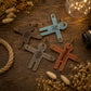 Four leather-shaped keychains arranged on a rustic wooden surface with dried flowers, rope, fairy lights, and a camera lens as styled props. The keychains are in brown, light blue, gray, and terracotta colors, each with engraved initials or names.