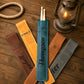 Five personalized leather drumstick pouches in brown, tan, blue, and gray, arranged on a rustic wooden table with a coiled rope and a lit lantern. The center blue pouch contains a pair of drumsticks and is personalized with the name 'Thompson'.