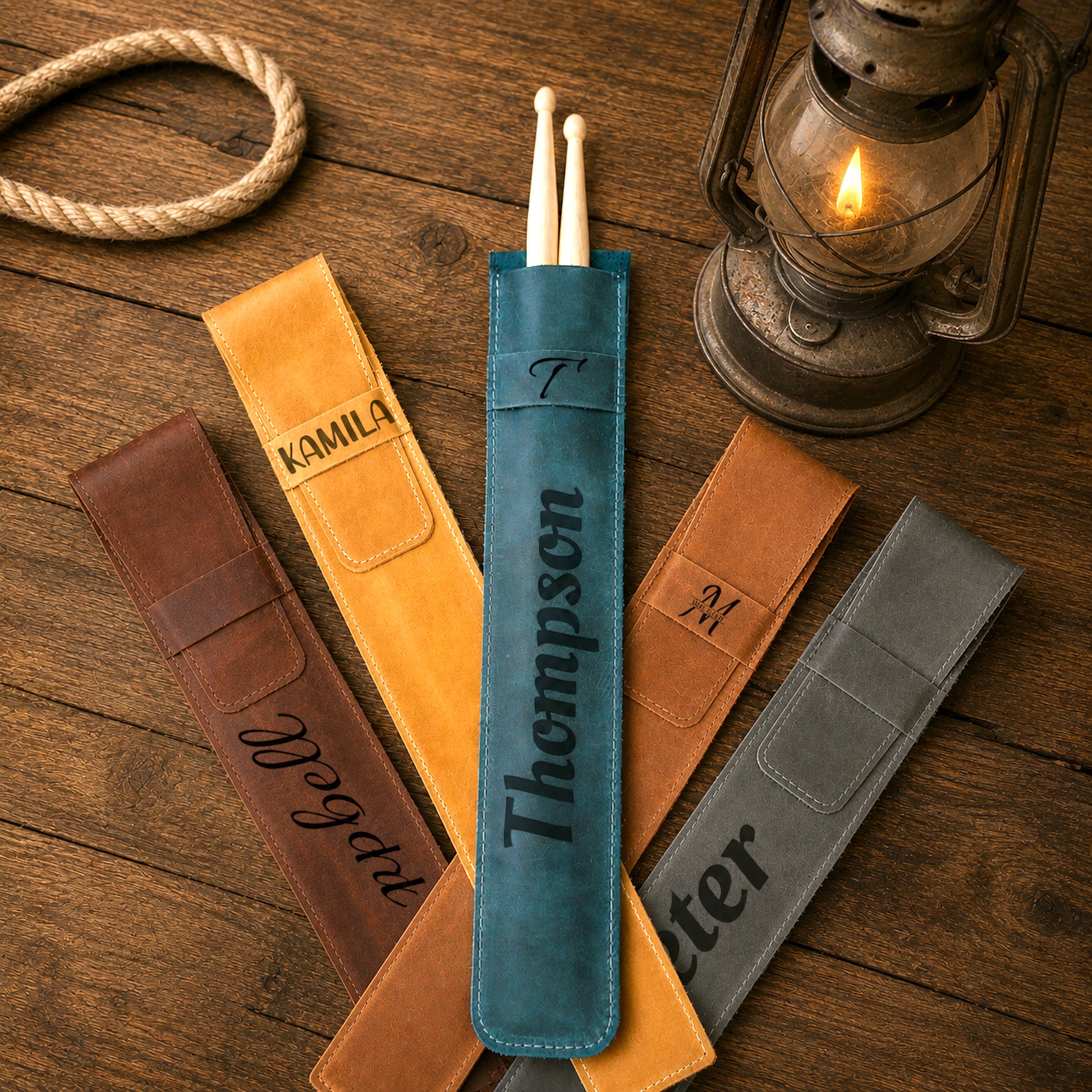 Five personalized leather drumstick pouches in brown, tan, blue, and gray, arranged on a rustic wooden table with a coiled rope and a lit lantern. The center blue pouch contains a pair of drumsticks and is personalized with the name 'Thompson'.