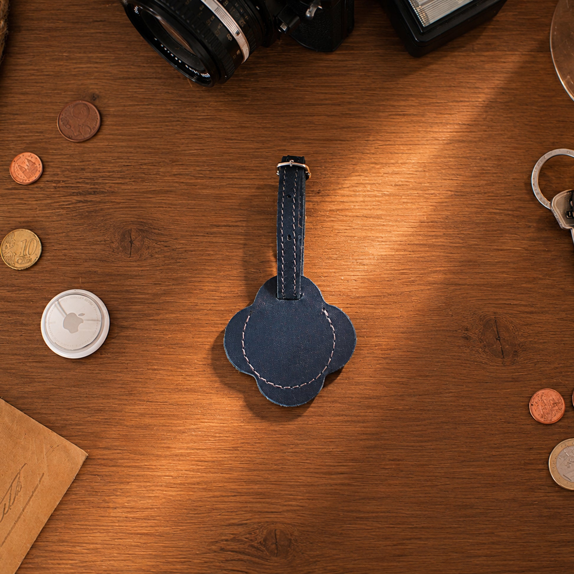 A dark blue leather luggage tag with a flower-shaped body and a matching strap, placed on a warm wooden tabletop surrounded by small props like coins, a camera, and keys.