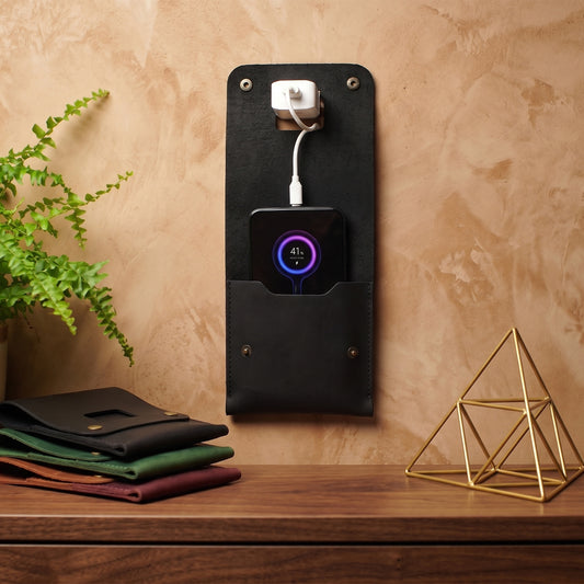 A black leather wall-mounted charging stand with a built-in USB outlet and a front pocket holding a phone, shown on a wooden table against a textured beige wall with decorative plants and geometric objects.