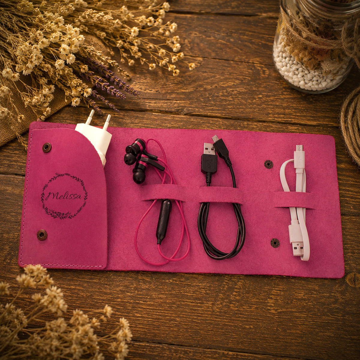 A pink felt travel charging pouch laid flat on a rustic wooden surface, holding a white wall plug, black wired earphones, a black USB cable, and a white USB-C cable. Dried flowers and decorative props surround the pouch.