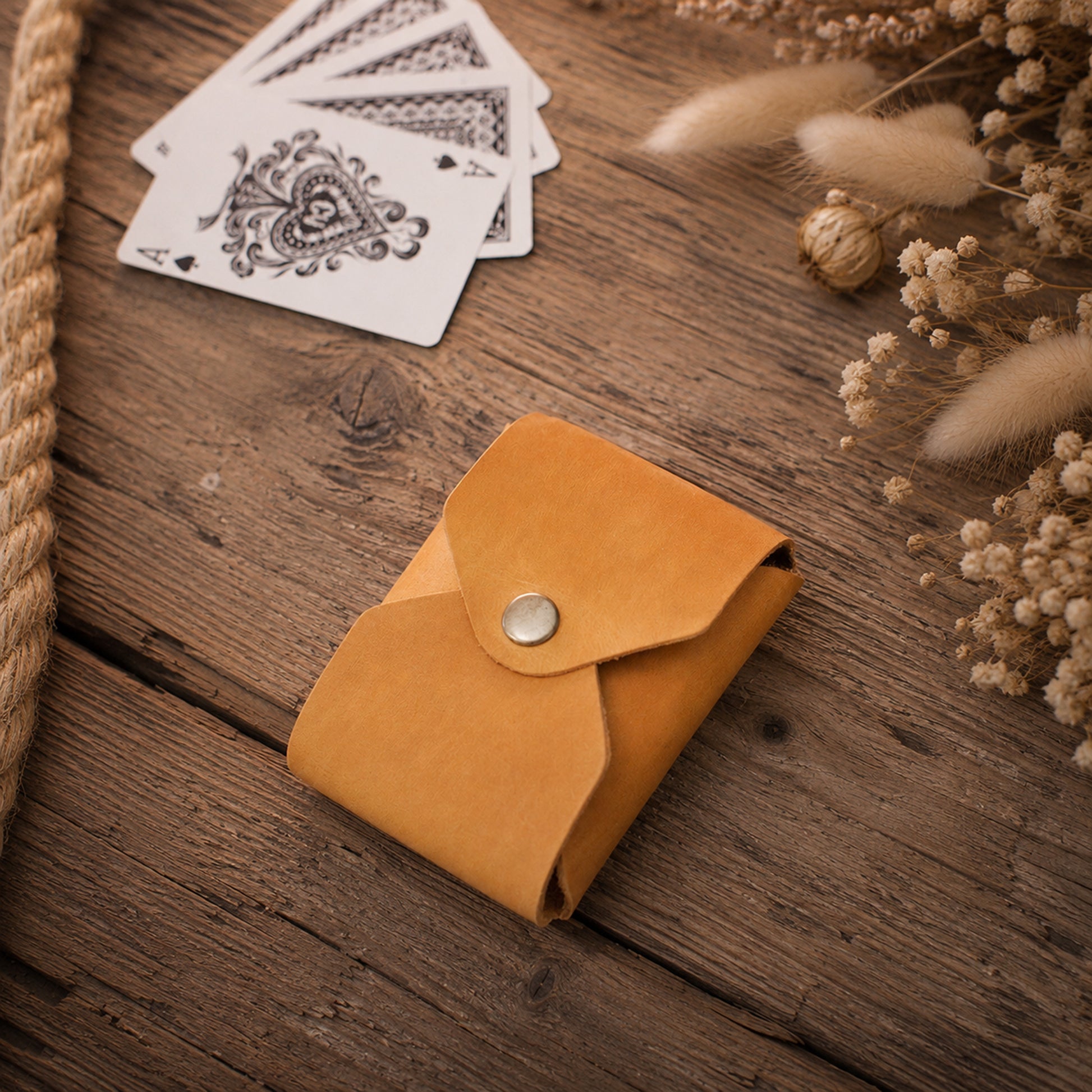 A small tan leather card holder or wallet with a snap button closure, placed on a rustic wooden surface with playing cards and dried flowers nearby.