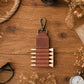 A brown leather pencil pouch with a metal clip, shown on a rustic wooden tabletop with dried flowers, glasses, and a pocket watch.