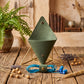 A green geometric cone-shaped wall hook or hanging ornament displayed on a gold metal stand on a rustic wooden table, with blue wired earbuds in a loop below and dried flowers and a potted fern in the background.