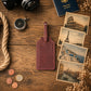 A dark reddish-brown leather luggage tag lies on a rustic wooden tabletop, surrounded by travel-themed props including a camera, compass, passport, coins, rope, dried flowers, and vintage-style Rome, Paris, Santorini, and Venice postcards.