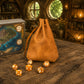 A tan leather drawstring pouch stands on a rustic wooden table, with several translucent amber polyhedral dice scattered in front. A fantasy map box is partially visible on the left, and the scene is set in a warmly lit medieval-style room with stone walls and round windows.