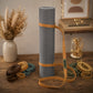 A gray textured yoga mat rolled up and secured with tan straps, standing on a wooden table. Several strap color variants are arranged around it, with dried flowers in a vase and warm string lights in the softly lit background.