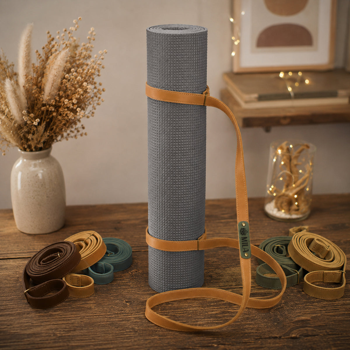 A gray textured yoga mat rolled up and secured with tan straps, standing on a wooden table. Several strap color variants are arranged around it, with dried flowers in a vase and warm string lights in the softly lit background.