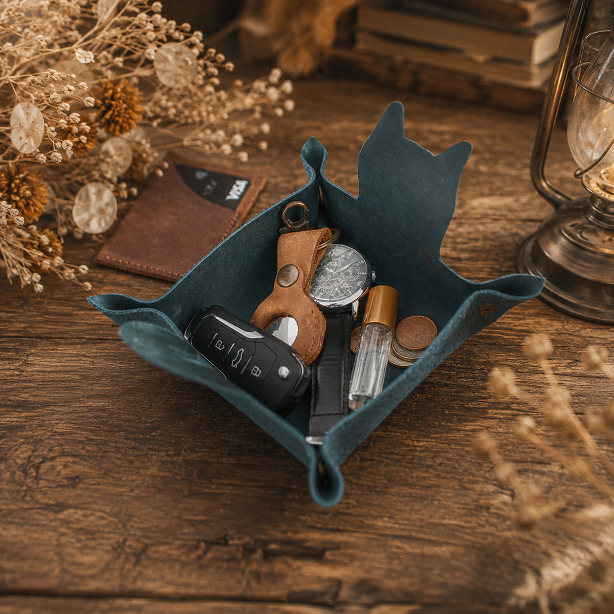 A teal blue felt car key holder tray filled with various keys, a watch, a perfume bottle, and coins, styled on a rustic wooden table with dried flowers and a vintage lantern.