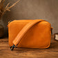 Orange leather crossbody bag on a wooden surface with a book and vase in the background.