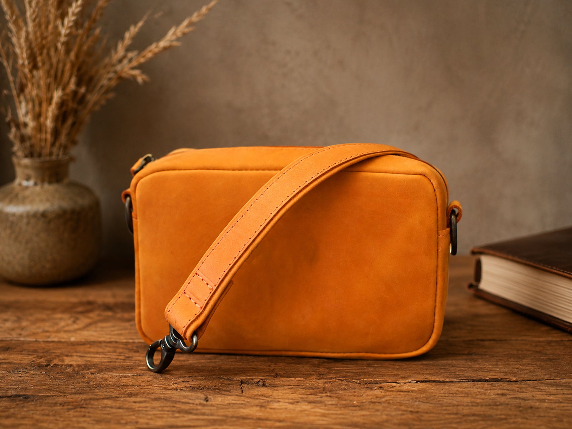 Orange leather crossbody bag on a wooden surface with a book and vase in the background.