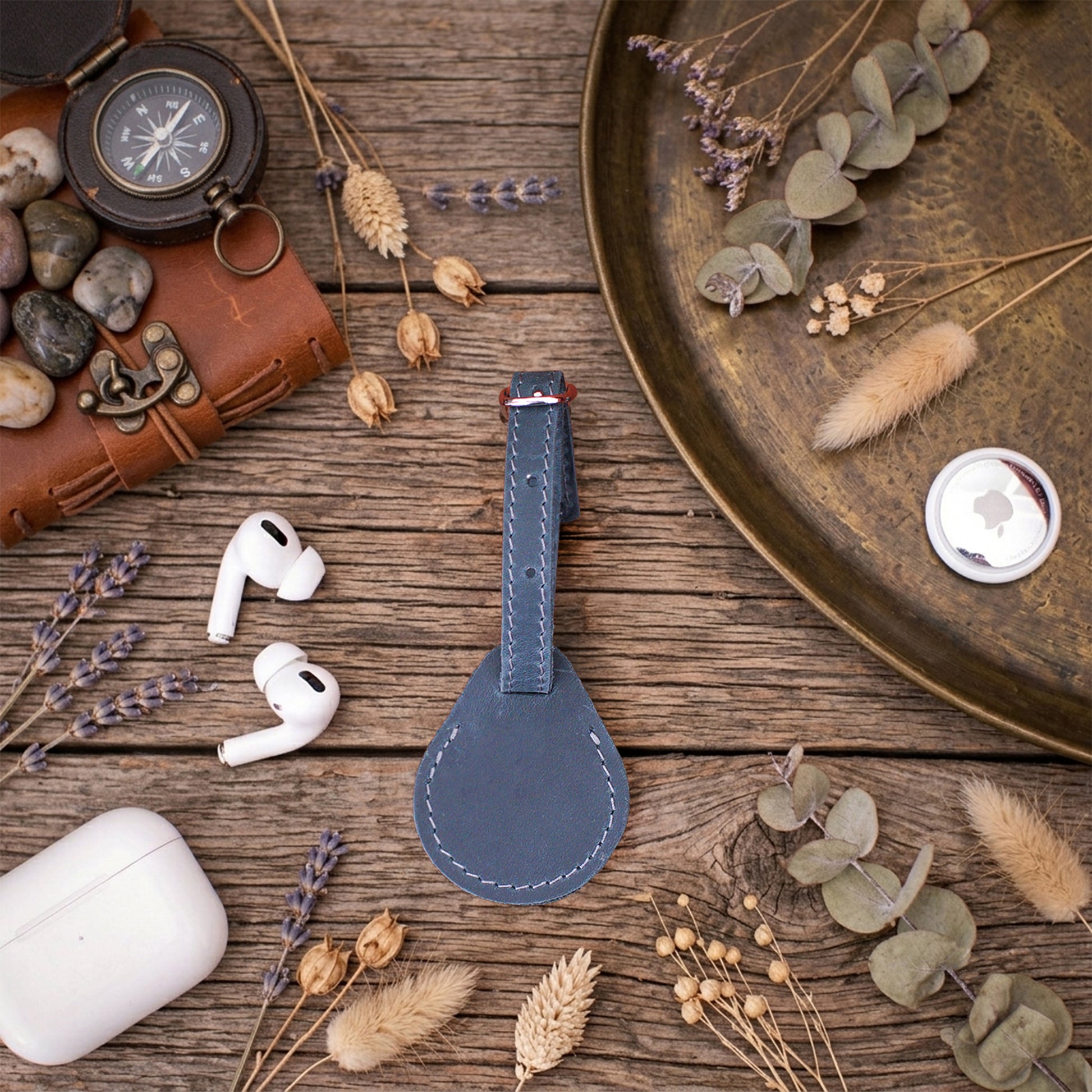 A dark blue leather luggage tag with white stitching is displayed on a rustic wooden table flat-lay. The scene is styled with travel-themed props including a brown leather case, a vintage compass, white wireless earbuds, a white charging case, and dried lavender and eucalyptus.