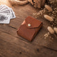 A small brown leather card holder wallet with a snap button closure, styled on a rustic wooden surface with playing cards and dried flowers nearby.