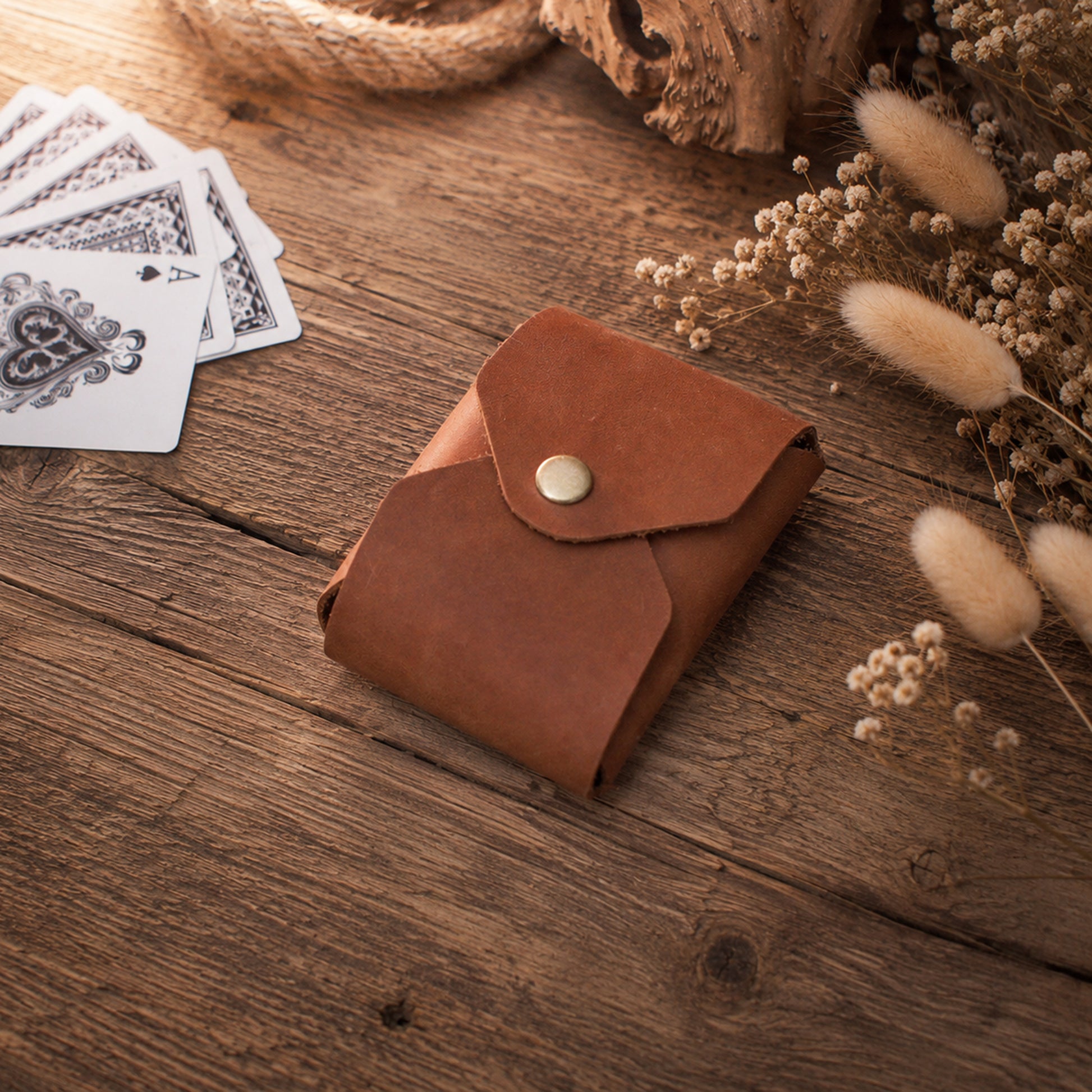 A small brown leather card holder wallet with a snap button closure, styled on a rustic wooden surface with playing cards and dried flowers nearby.