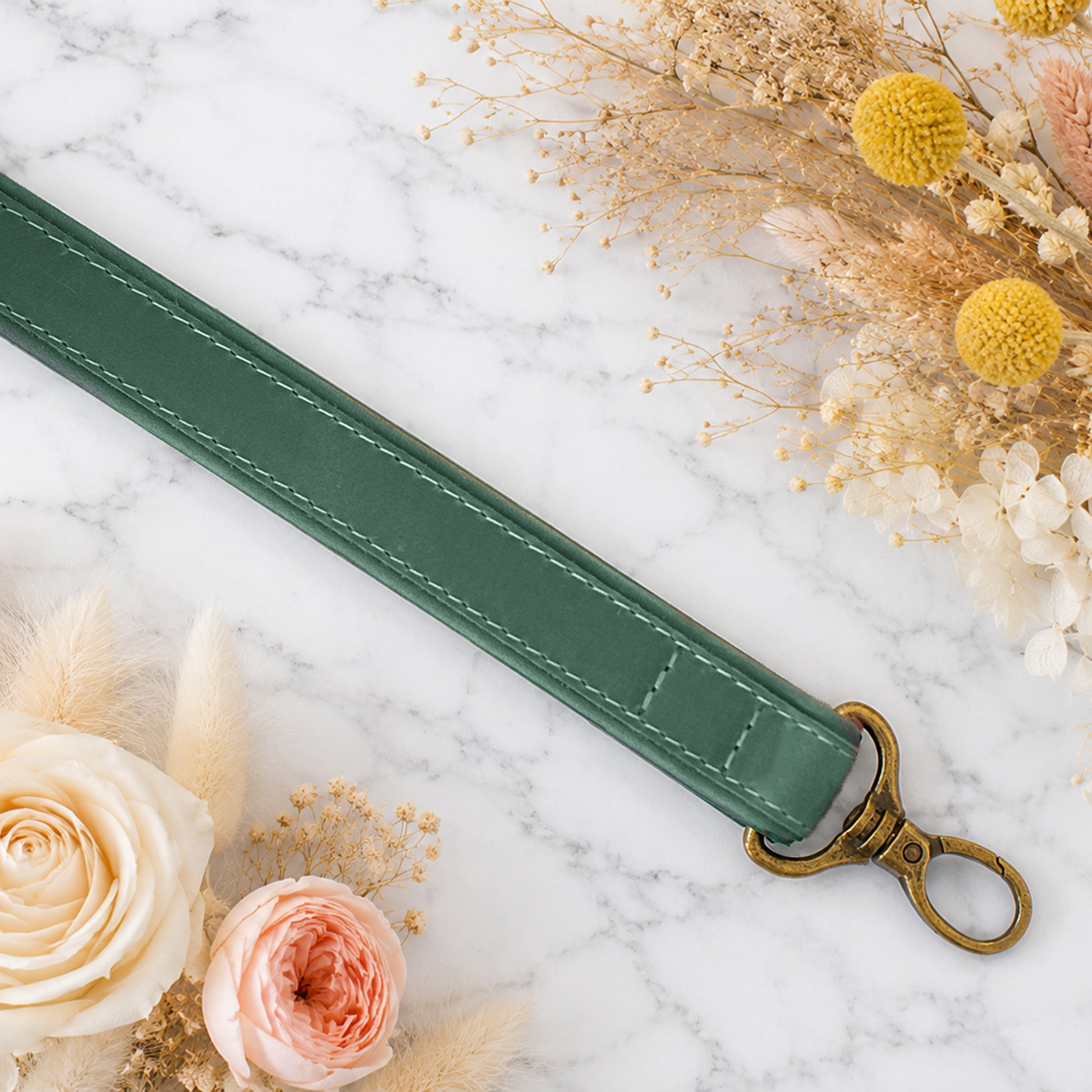 Green leather keychain with brass hook on a marble surface with flowers and dried plants.