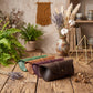 Four leather eyeglasses cases in green, tan, mauve, and dark brown are arranged on a rustic wooden table, styled with dried lavender, cotton, a macrame wall hanging, and a ceramic vase in the background.