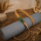 A rolled gray yoga mat with a tan leather carrying strap and a green brand tag reading 'Ridder', displayed on a wooden tabletop with dried floral decor in the background.