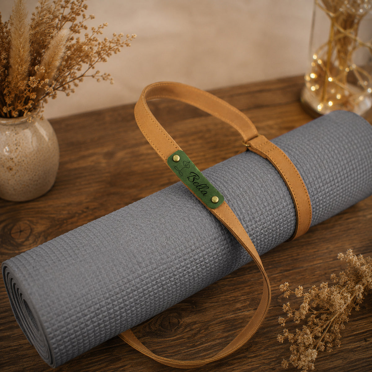 A rolled gray yoga mat with a tan leather carrying strap and a green brand tag reading 'Ridder', displayed on a wooden tabletop with dried floral decor in the background.