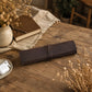 A dark brown leather journal or notebook cover wrapped with a matching tie strap, resting on a rustic wooden table surrounded by dried flowers, a book, and a small dish with earbuds.