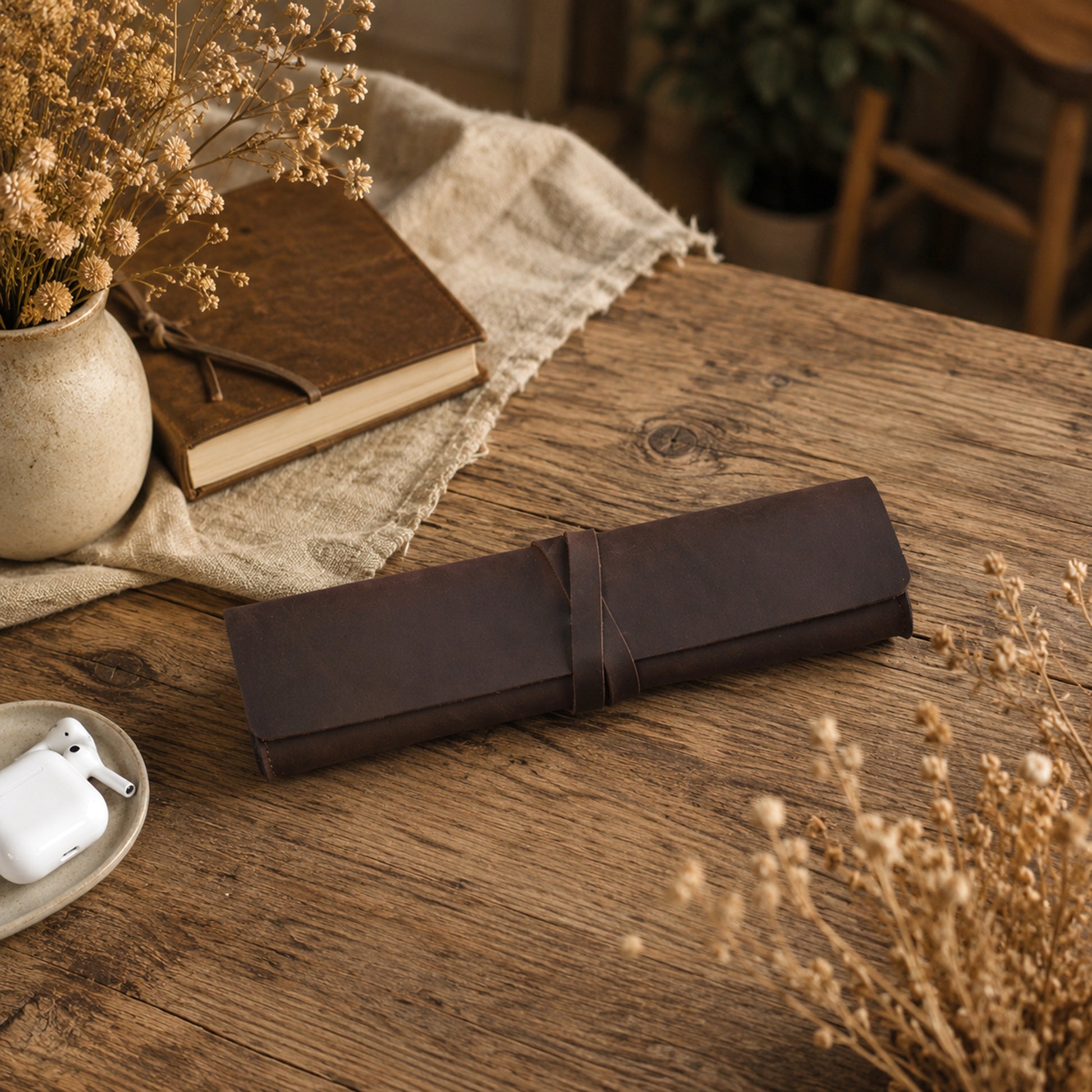 A dark brown leather journal or notebook cover wrapped with a matching tie strap, resting on a rustic wooden table surrounded by dried flowers, a book, and a small dish with earbuds.
