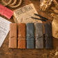 Five personalized leather journal covers in tan, brown, slate blue, gray, and charcoal, arranged on a rustic wooden table with vintage stationery props.