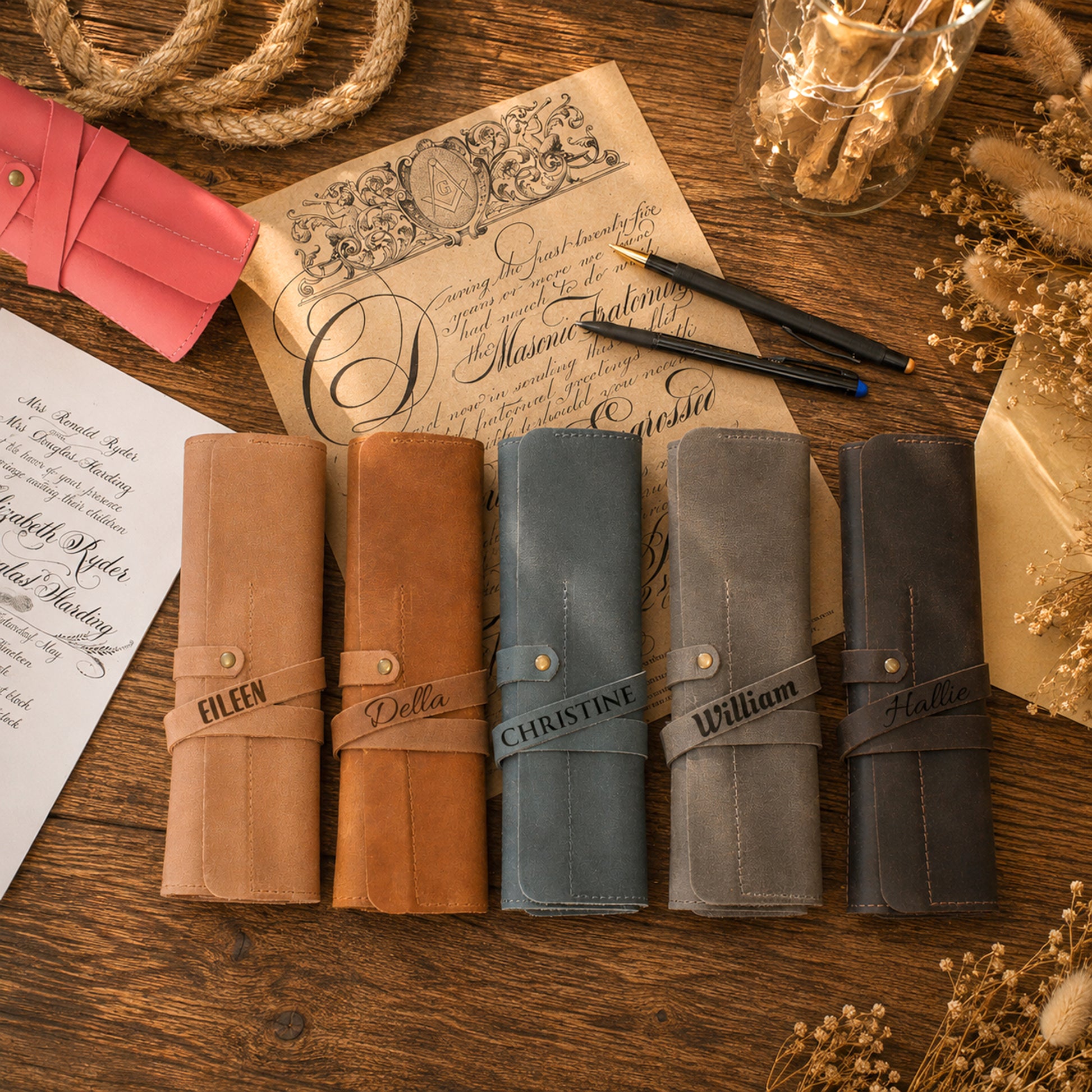 Five personalized leather journal covers in tan, brown, slate blue, gray, and charcoal, arranged on a rustic wooden table with vintage stationery props.