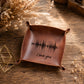 A square brown leather tray with rolled corners, featuring a black soundwave graphic and the words 'I love you' engraved on the bottom. The tray is displayed on a rustic wooden table with dried flowers and a small gold geometric prop.