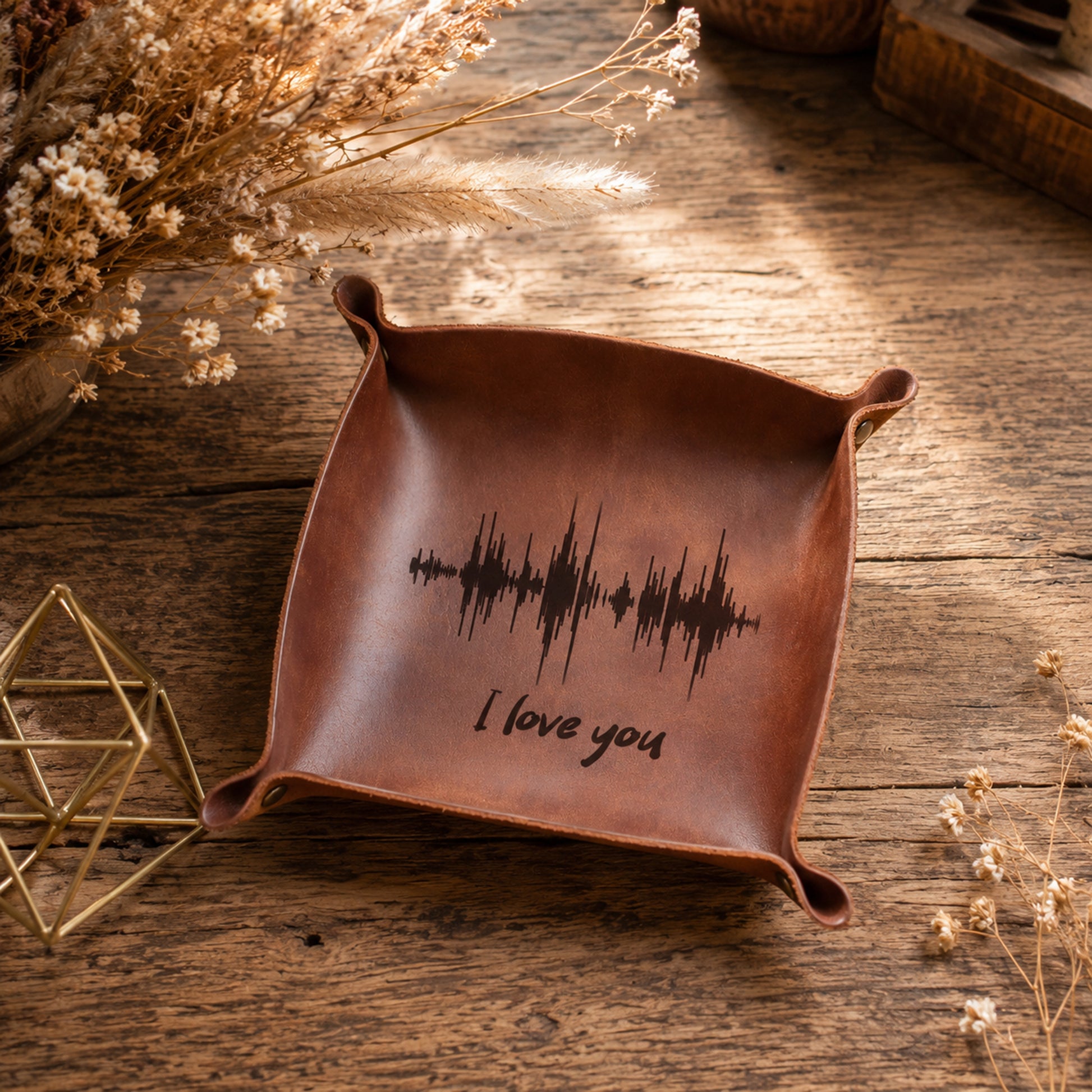 A square brown leather tray with rolled corners, featuring a black soundwave graphic and the words 'I love you' engraved on the bottom. The tray is displayed on a rustic wooden table with dried flowers and a small gold geometric prop.