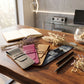 Four personalized leather card cases in gray, pink, tan, and navy are arranged on a wooden desk alongside dried flowers, a pen, and an open notebook in a modern office setting.