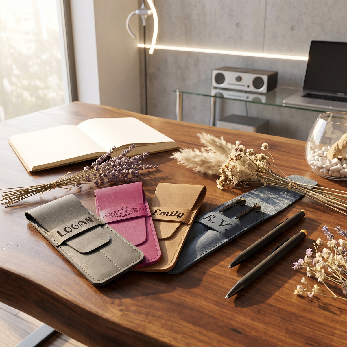Four personalized leather card cases in gray, pink, tan, and navy are arranged on a wooden desk alongside dried flowers, a pen, and an open notebook in a modern office setting.