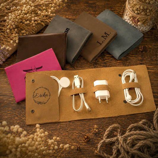A tan roll-up organizer case laid open on a wooden surface, holding a white charging cable, a white power adapter, and a white magnetic charging pad. Several closed matching cases in brown, gray, blue, and pink are arranged behind it, with decorative dried flowers and rope around the scene.