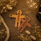 Two personalized leather keychains shaped like children, one tan and one brown, engraved with the names 'DANIEL' and 'SABRINE' respectively, displayed on a rustic wooden surface with dried flowers and warm string lights.
