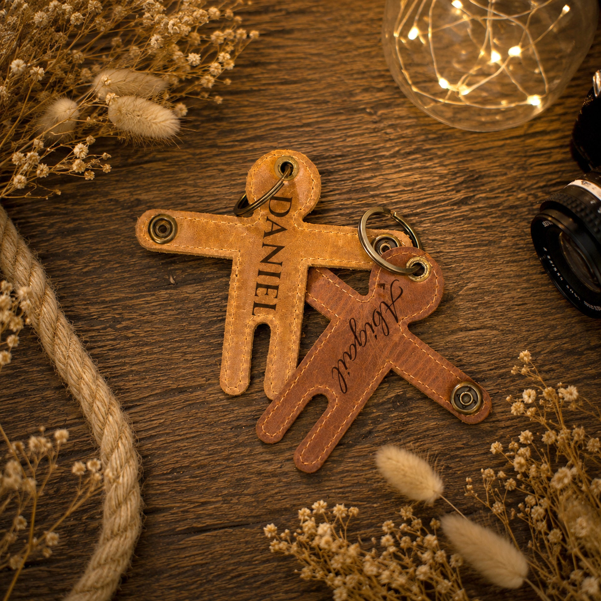 Two personalized leather keychains shaped like children, one tan and one brown, engraved with the names 'DANIEL' and 'SABRINE' respectively, displayed on a rustic wooden surface with dried flowers and warm string lights.