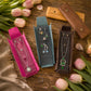 Three velvet jewelry organizer sleeves in pink, teal, and brown, each holding a necklace and earrings, arranged on a wooden table with tulips and rings.