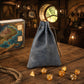 A dusty blue drawstring pouch bag standing on a rustic wooden table, with amber-colored polyhedral gaming dice scattered around it and a fantasy map box in the background.