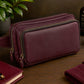 Maroon leather crossbody bag on a wooden surface with a plant, bottle, and candle in the background.