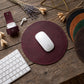 A round burgundy leather mouse pad on a rustic wooden desk, styled with a white wireless mouse, a matching burgundy leather sleeve, a keyboard, dried flowers, and other color options of the mouse pad visible on the right.