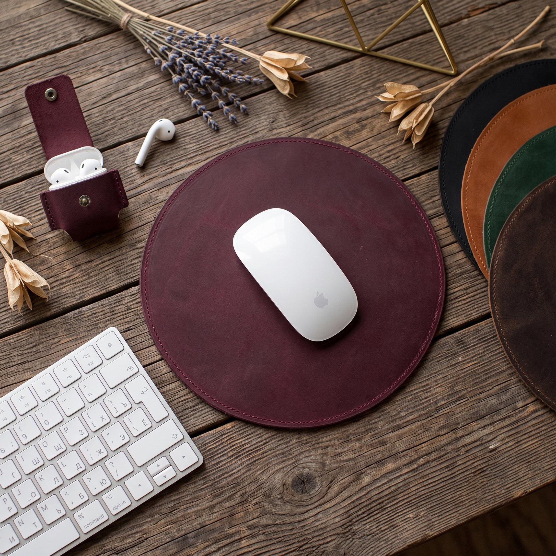 A round burgundy leather mouse pad on a rustic wooden desk, styled with a white wireless mouse, a matching burgundy leather sleeve, a keyboard, dried flowers, and other color options of the mouse pad visible on the right.