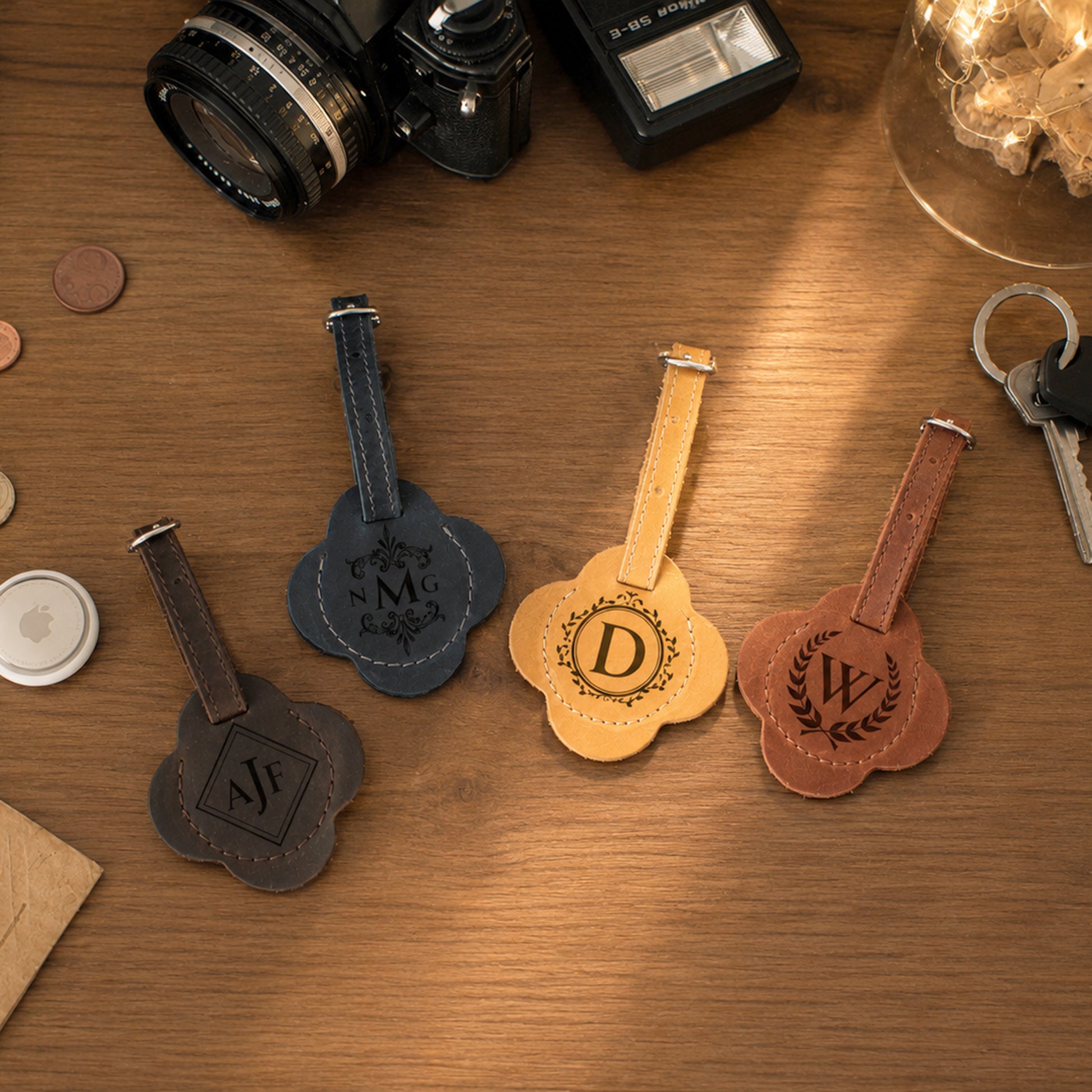 Four personalized leather luggage tags in different colors (brown, navy, tan, and reddish-brown) arranged on a wooden table with vintage props like a camera, keys, and a lamp.