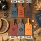 Six personalized leather luggage tags in blue, tan, brown, red, gray, and yellow, arranged on a rustic wooden background with travel-themed props. Text labels above and below indicate the available colors: DENIM, TAWNY, CHOCOLATE, BROWN, GRAY, and YELLOW.