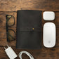Black leather cable organizer, glasses, white Apple mouse, and charger on a wooden surface