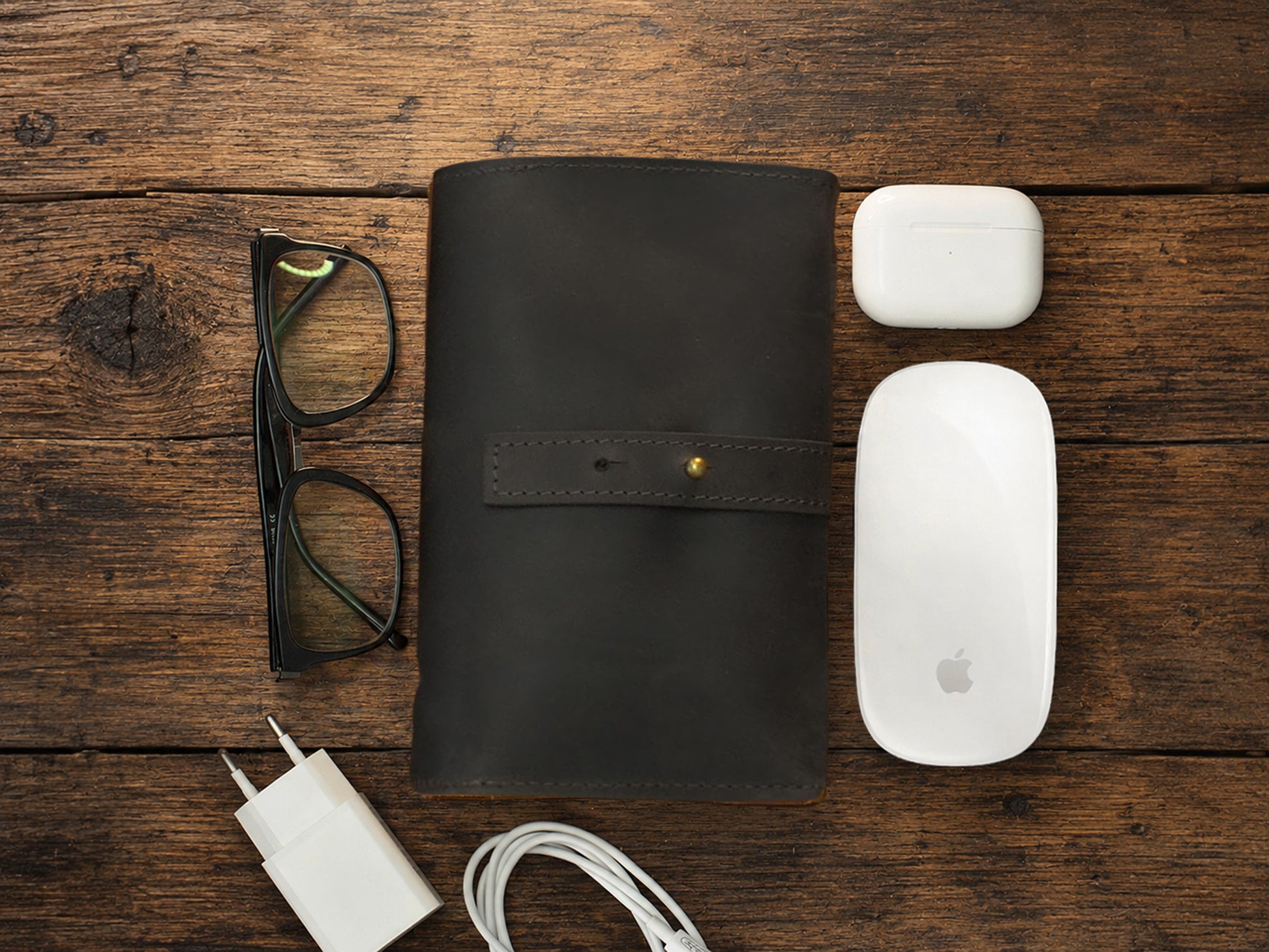 Black leather cable organizer, glasses, white Apple mouse, and charger on a wooden surface