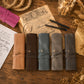Five personalized leather journal covers in tan, brown, blue-gray, and charcoal are arranged on a rustic wooden table with vintage-style papers, pens, rope, and dried flowers. The covers are shown closed with leather straps and brass snap buttons, each personalized with a name.