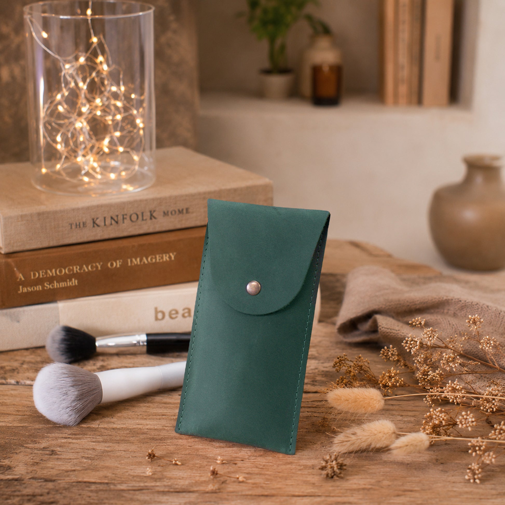 A dark teal green leatherette pouch with a silver snap button, standing on a rustic wooden table surrounded by makeup brushes, dried flowers, and decorative books.