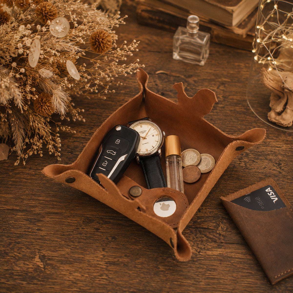 A brown leather tray organizer styled on a wooden table, holding a car key fob, watch, perfume bottle, coins, and a small round accessory. A brown leather wallet is placed beside it, with dried flowers, books, and decorative lights in the background.