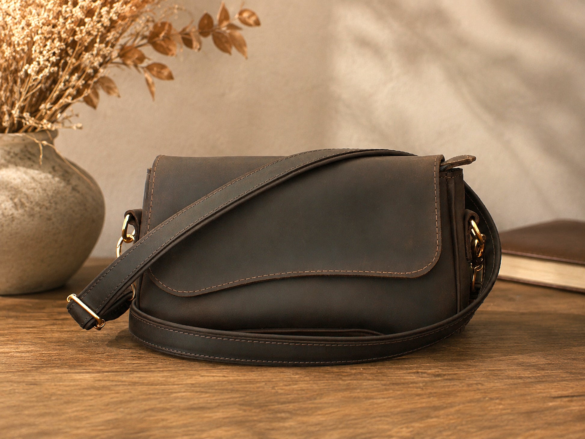 Brown leather bag on a wooden surface with a vase of dried flowers in the background