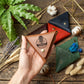 A hand holds a triangular brown leather card holder with an engraved logo that reads 'HENRY' and a guitar graphic. Surrounding it are other folded leather card holders in red, navy, and green, along with decorative dried grasses, cotton bolls, a fern, and a gold geometric frame on a rustic wooden surface.
